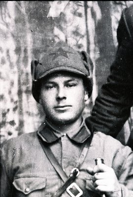 Close-up portrait of a young man in a military uniform and peaked cap. Features visible red armband and belt with buckle. The...