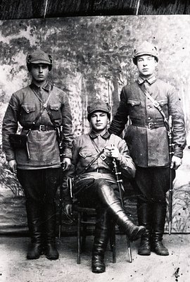 Three men in military uniforms with peaked caps and high boots pose for a studio portrait. Two stand flanking a seated man wi...