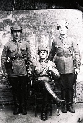 Three men in military uniforms pose for a studio portrait. Two stand, one seated; each wears a cap, belt, and boots. The seat...