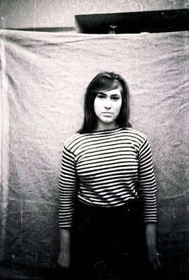 Striking monochrome portrait of a young woman with a direct gaze, wearing a striped long-sleeved shirt and dark skirt. Likely...