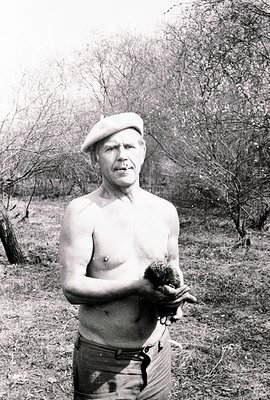 A man, bare-chested and wearing a beret, stands outdoors holding a hedgehog. Visible foliage suggests a wooded area. The imag...