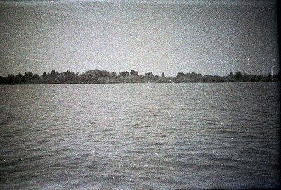A wide, monochrome landscape featuring a still body of water extending to a dense treeline. Strong grain and visual artifacts...