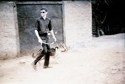 A man walks two dogs along a gravel path outside a building with a large, weathered wooden door. The man wears dark clothing ...