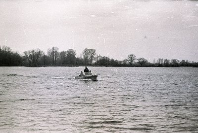 A small motorboat with two figures travels across a calm body of water. Bare trees line the far bank, suggesting early spring...