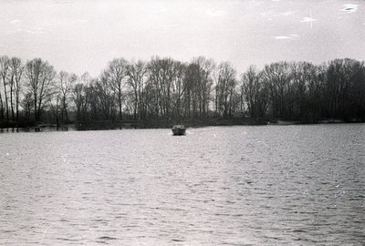 Monochromatic view of a calm body of water, likely a lake or wide river. Dense, leafless trees line the distant shore. A smal...
