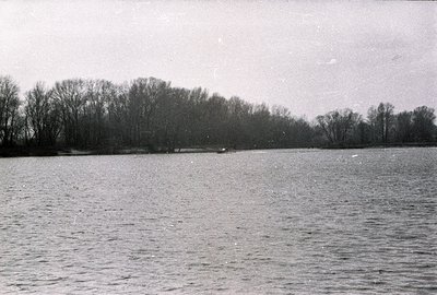 A wide, still body of water fills the frame, reflecting a distant treeline under a muted sky. A small boat is visible mid-sce...