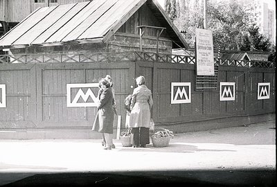 Two women in traditional clothing carry baskets along a street lined with a wooden fence and signage featuring a stylized "M"...