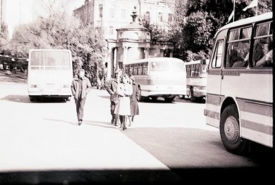 Three figures, two adults and a child, walk along a paved road lined with buses. A distinctive pavilion structure is visible ...