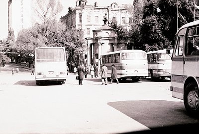 A black and white photograph depicts a street scene, likely in Bulgaria. Three buses are visible, partially obscuring a grand...