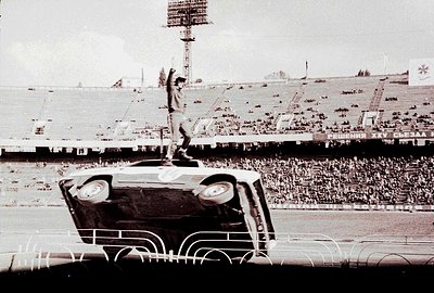 A lone man, seemingly a performer or athlete, stands triumphantly atop an overturned car during an outdoor stadium event. The...