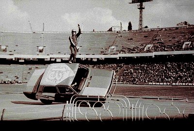 A man stands atop a "Burev" sedan, raising his arm in a celebratory gesture. The vehicle is positioned on a track within a la...