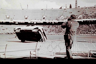 A dramatic scene: a car overturned on a track during a motorsport event, viewed from behind a figure in a suit. The blurred b...