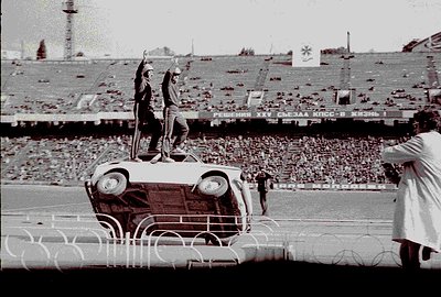 A man stands atop a vintage Volga sedan at a stadium, raising his arm in a gesture that could be interpreted as a salute. The...