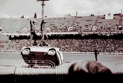 A daredevil in a jumpsuit stands atop an overturned vehicle amidst a large crowd at a stadium. Cyrillic text on banners and s...