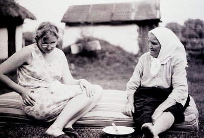 Two women sit on a mattress outdoors. One, in a patterned tank top and glasses, gestures while the other, wearing a headscarf...