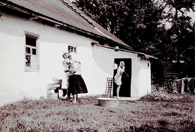 A black and white photo depicts two women near a modest, whitewashed cottage with a low-slung roof. One woman stands outside,...