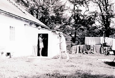Exterior view of a small, whitewashed building with a single person framed within the doorway. Another person, seemingly a ch...