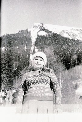 Young woman in a fair isle sweater and pleated skirt stands in a snowy, forested landscape with a prominent ski slope visible...