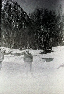 A young person bundled in a thick sweater & hat stands in a snow-covered landscape. A steep, rocky mountain looms in the back...