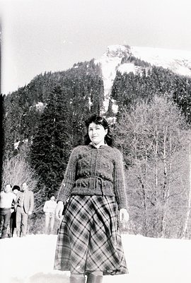 A young woman stands in a snow-covered landscape, wearing a cable-knit sweater and a plaid skirt. A group of people in winter...