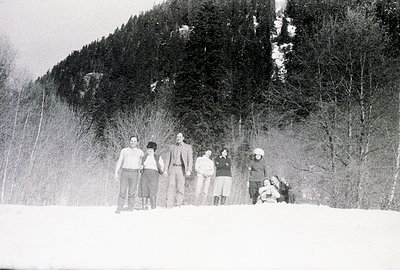 Group portrait of six individuals in winter clothing, standing on a snowy field bordered by a dense evergreen forest. Appears...