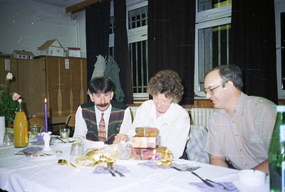 Three individuals seated around a formally set table with a wrapped gift centerpiece. Men's attire and interior design sugges...