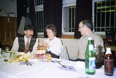 A candid moment around a table, three individuals appear to be opening gifts. Man in a patterned vest and mustache sits left....