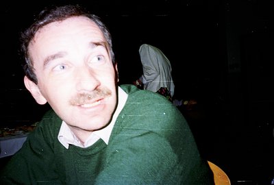 A man with a mustache and wide eyes is captured in a close-up, likely a candid moment. He wears a green sweater over a white ...