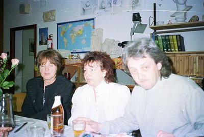 Three people seated at a table laden with drinks, appearing to be engaged in conversation. Visible are a world map, a bust sc...