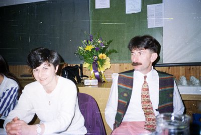 Two individuals are seated indoors, likely at an event or meeting. Woman in white shirt; man in patterned vest and tie. A vib...