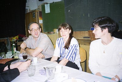 Three individuals seated at a table covered with patterned linens and glassware. A man in glasses wears a striped shirt; two ...