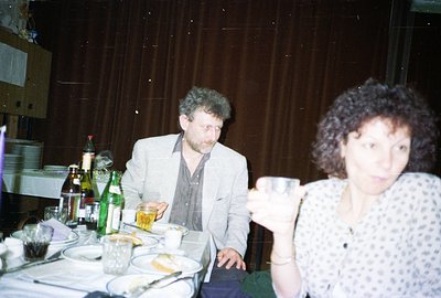 A seated couple shares a table laden with food and drink. The man, wearing a grey blazer, looks downward. The woman, with vol...