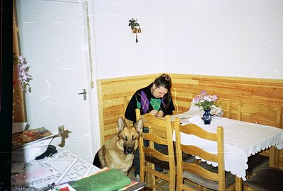 A young woman sits writing in a wooden-paneled room, a German Shepherd dog resting near her. Table draped with lace; floral a...