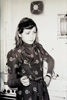 A woman in a floral patterned dress and headband poses indoors, likely 1970s. She holds a film reel spool, hinting at a conne...