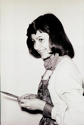 A smiling woman with a short, dark bob haircut is captured in a posed studio portrait. She wears a patterned paisley top and ...