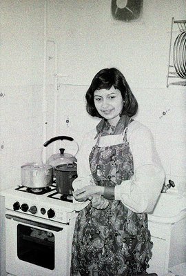 A woman stands in a kitchen, smiling. She wears a patterned dress and apron, with a patterned scarf. The kitchen features a v...