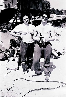 Two individuals, a man and a woman, pose outdoors. The man, shirtless, holds a bottle; the woman wears a white top and trouse...