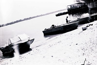 A black and white image captures two small motorboats resting on a sandy shoreline beside a river. A lone man is positioned i...