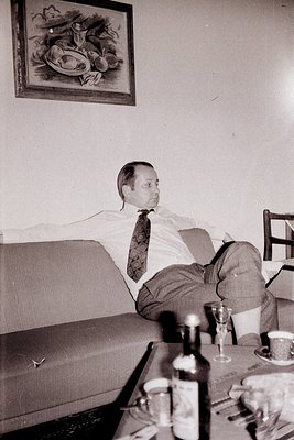 Man in suit and tie reclines on a mid-century modern sofa. A still life painting hangs behind. Table with drinks and ashtrays...