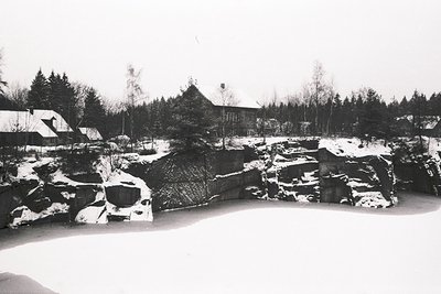 Striking monochrome landscape depicting a frozen quarry lake and surrounding rock formations. Sparse trees and a distant buil...