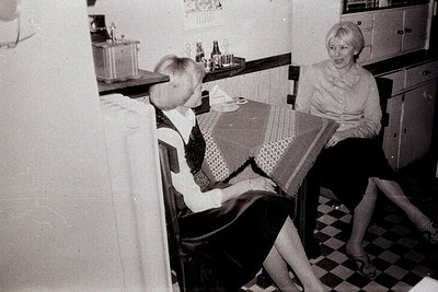 Two women seated at a table in a kitchen, engaged in conversation. The kitchen features checkered flooring, dark wood cabinet...