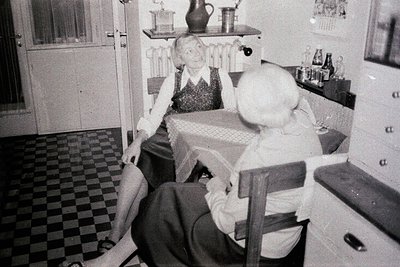 A seated woman in a skirt and blouse is captured in a domestic interior. Checkered tile flooring and a patterned tablecloth d...