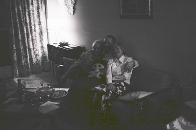 A man embraces a woman seated on a patterned sofa within a dimly lit room. A stuffed bear rests between them. Visible furnish...