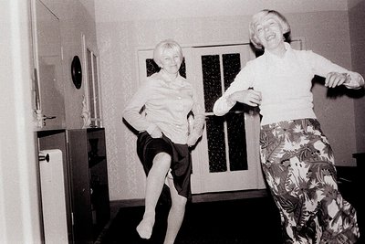 Two women in patterned and dark skirts, tights, and vintage blouses are playfully leaping in a home interior. Black and white...