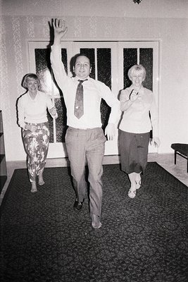 A man in a suit and tie raises his arm, seemingly greeting someone off-camera. Two women, one in patterned pants and one in a...