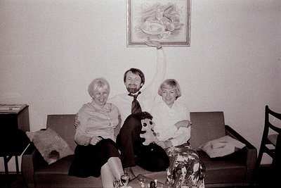 A candid black-and-white photograph captures a family moment. Two women and a man are seated on a sofa, a baby held aloft by ...