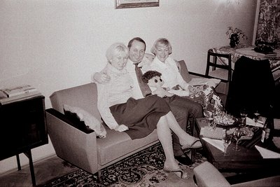A family portrait captures a man and two women seated on a sofa alongside a doll. The decor suggests a 1960s or 70s interior ...