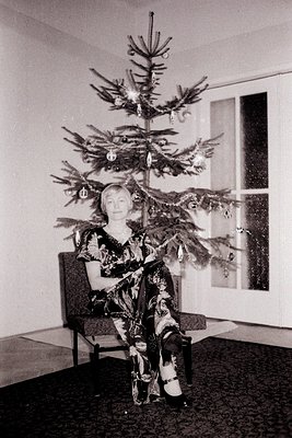 A woman in a patterned dress and Mary Jane shoes sits elegantly in a chair before a decorated Christmas tree. The scene, like...