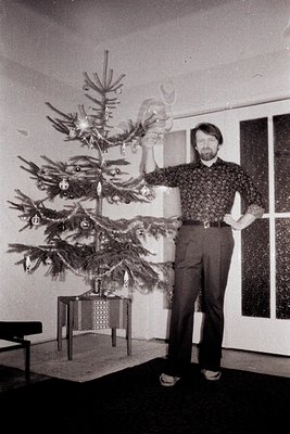 Man in 1970s attire stands proudly beside a decorated Christmas tree. Note the patterned shirt, high-waisted trousers, and pl...