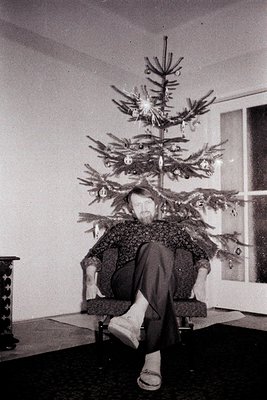 A man with a full beard playfully poses behind a decorated Christmas tree, seated in a dark upholstered armchair. He wears st...